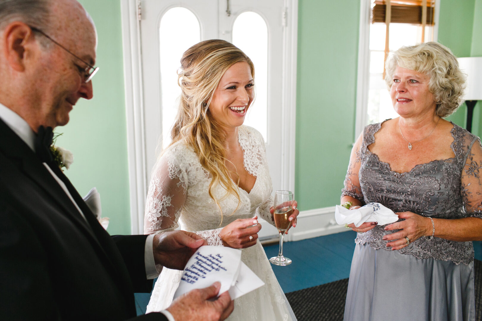 A bride in a white dress smiles, holding a glass at Willow Creek Winery, standing between two older adults in formal attire, each clutching handkerchiefs. They are inside a room with green walls and white doors.