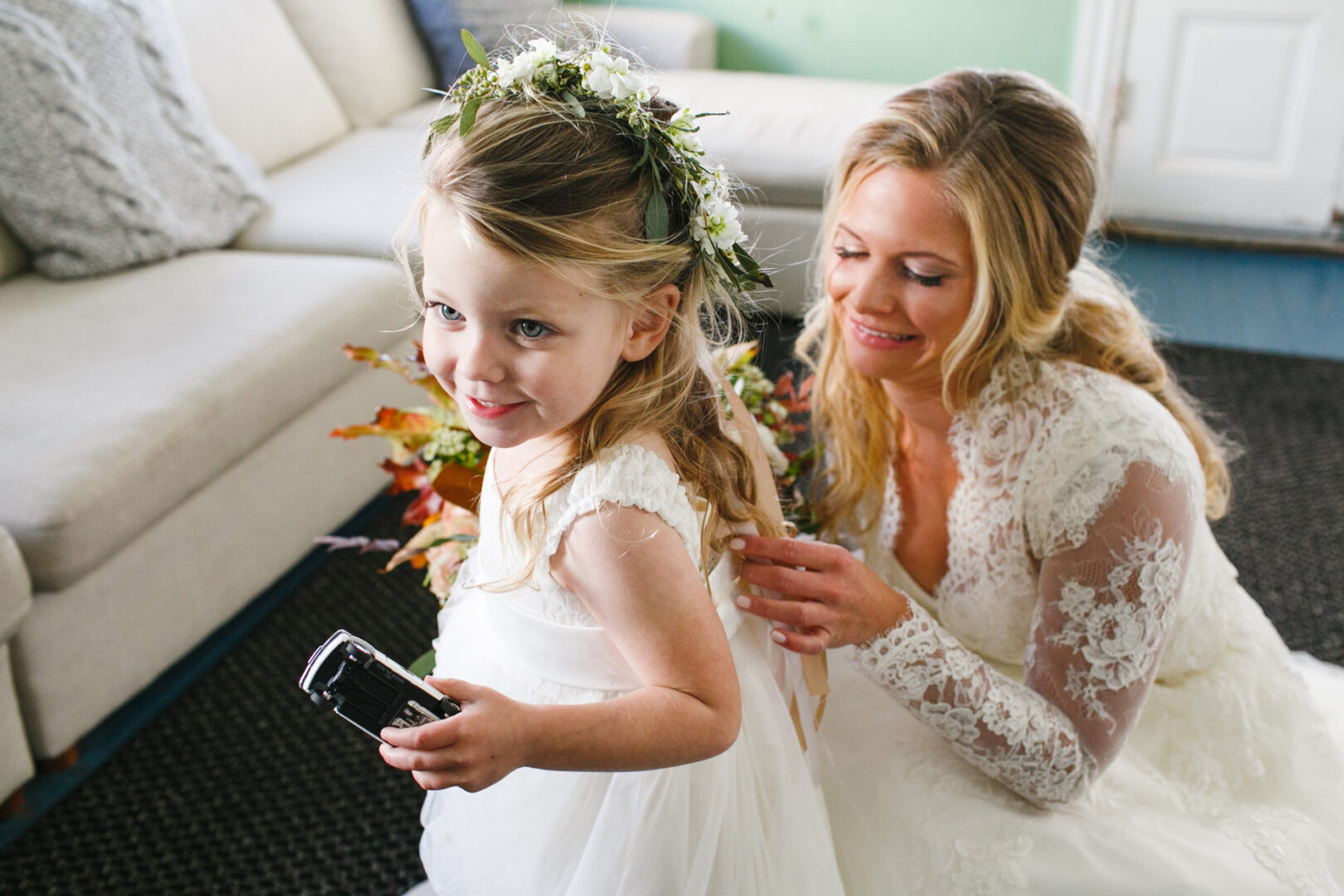 At Willow Creek Winery, a bride in a lace dress gently adjusts the flower crown of a young flower girl, who clutches a camera ready to capture every magical moment.