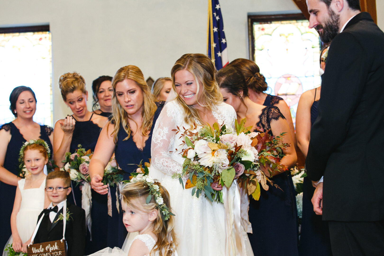 In a picturesque wedding scene at Willow Creek Winery, the bride stands gracefully in her white dress, clutching flowers. She's surrounded by bridesmaids, children, and the groom. The group gathers indoors beneath stained glass windows and near an American flag.