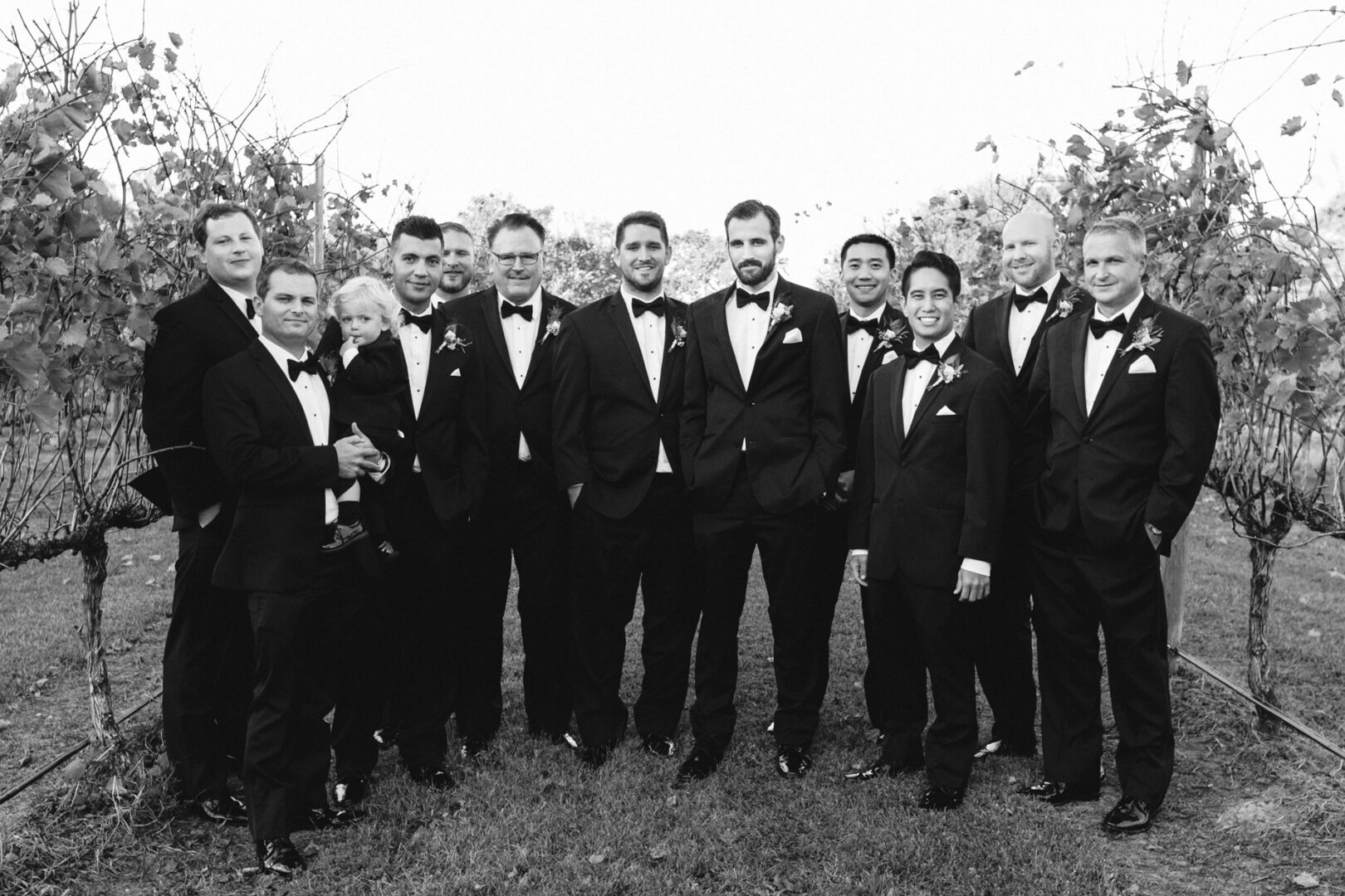 A group of men in tuxedos stand together outdoors in front of the vineyard rows at Willow Creek Winery, posing for a formal photo.