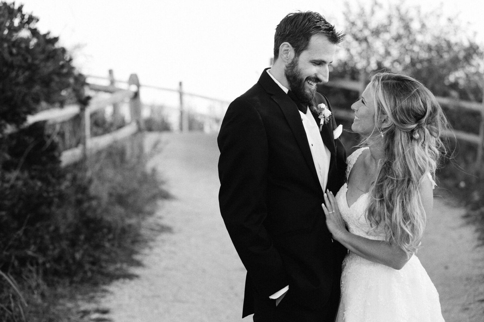 A bride and groom in formal attire stand together on a path at Willow Creek Winery, smiling at each other. The background features a wooden fence and lush foliage. The image is beautifully captured in black and white.