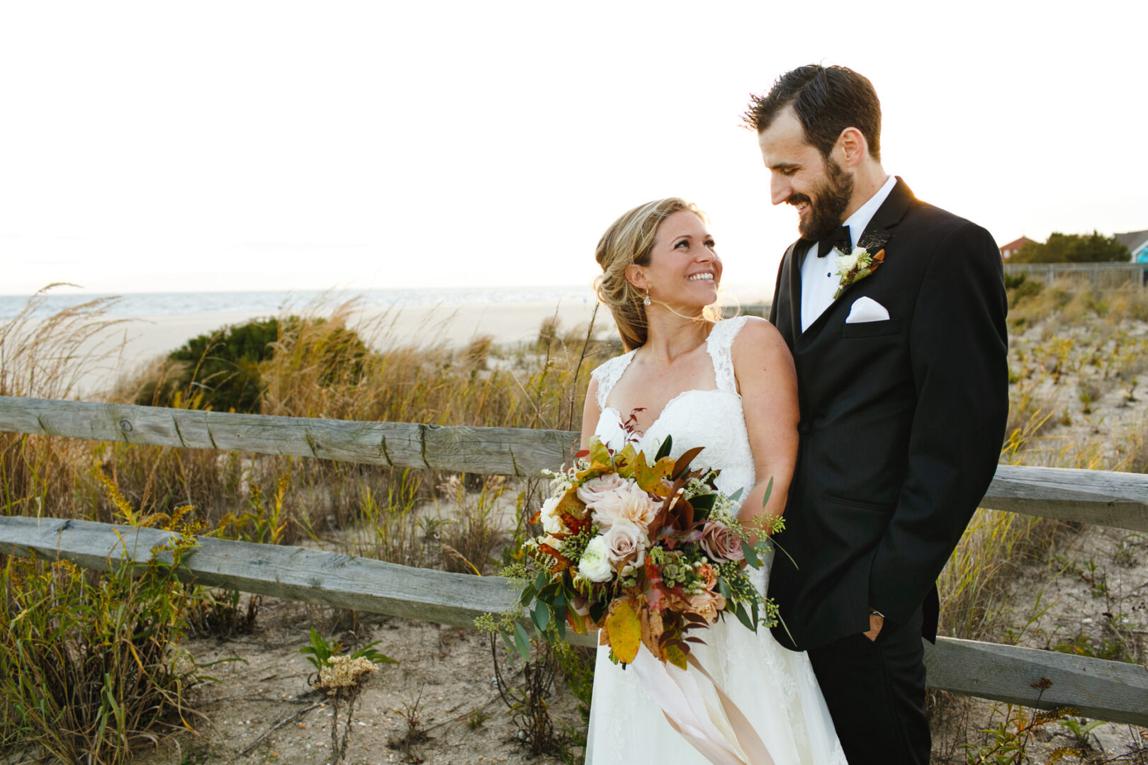Bride and groom smiling at each other in front of a wooden fence, with the beach and grassy dunes at Willow Creek Winery in the background. The bride holds a bouquet.