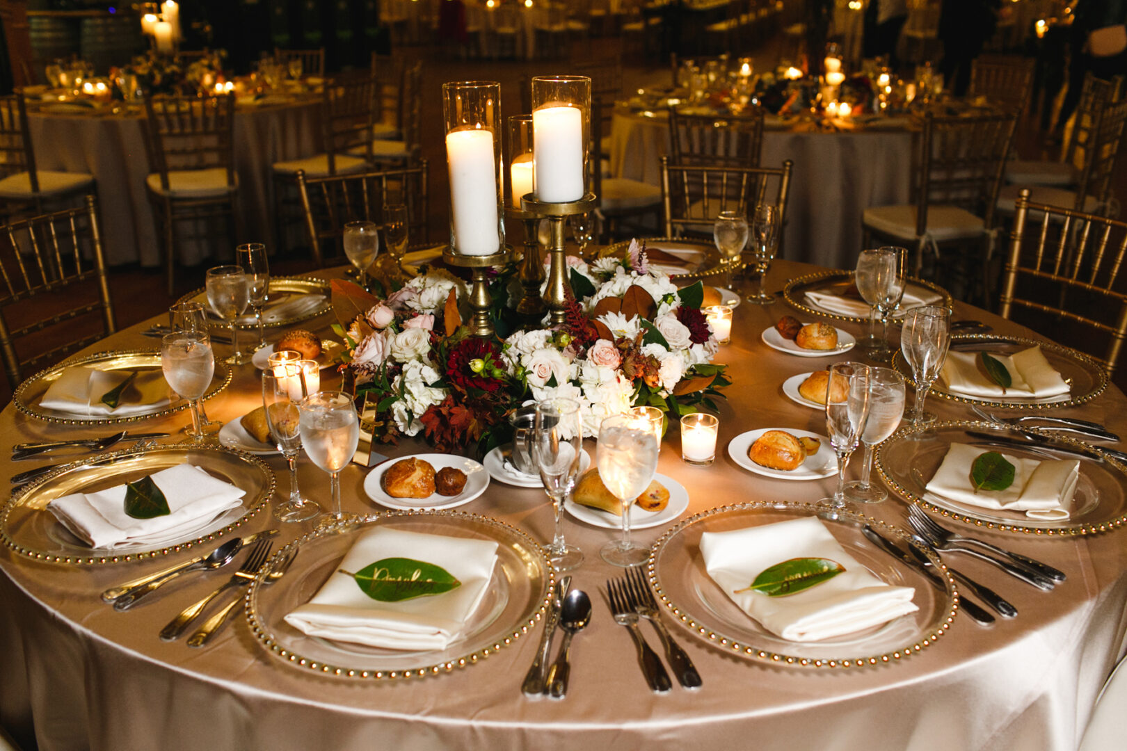 Elegant table setting featuring a floral centerpiece, candles, and clear plates is reminiscent of an evening at Willow Creek Winery. Each place boasts fine silverware, glassware, a napkin adorned with a leaf, and small bread rolls beside the plates.