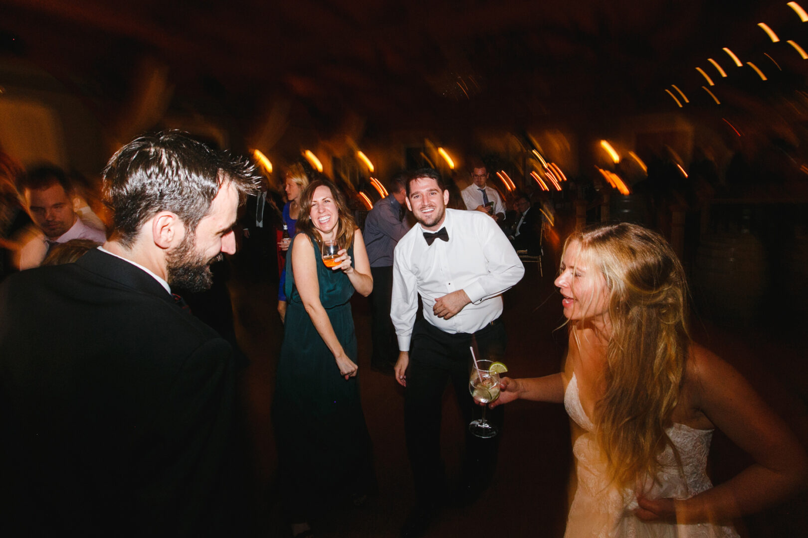 At Willow Creek Winery, people dance joyfully at a lively event; a woman in a white dress and a man in a bow tie mingle with others, all smiling and holding drinks. Warm lighting and blurred motion capture the winery's energetic vibe.