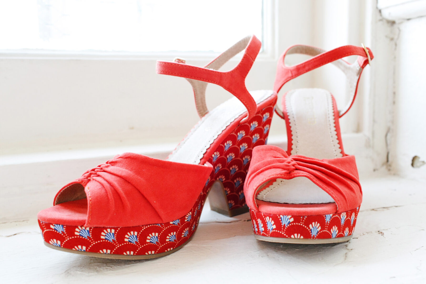 Red high-heeled sandals with ankle straps, featuring decorative blue and white floral patterns on the platform and heel, elegantly await their moment on a white wooden surface—perfect for a Baldwin School wedding.