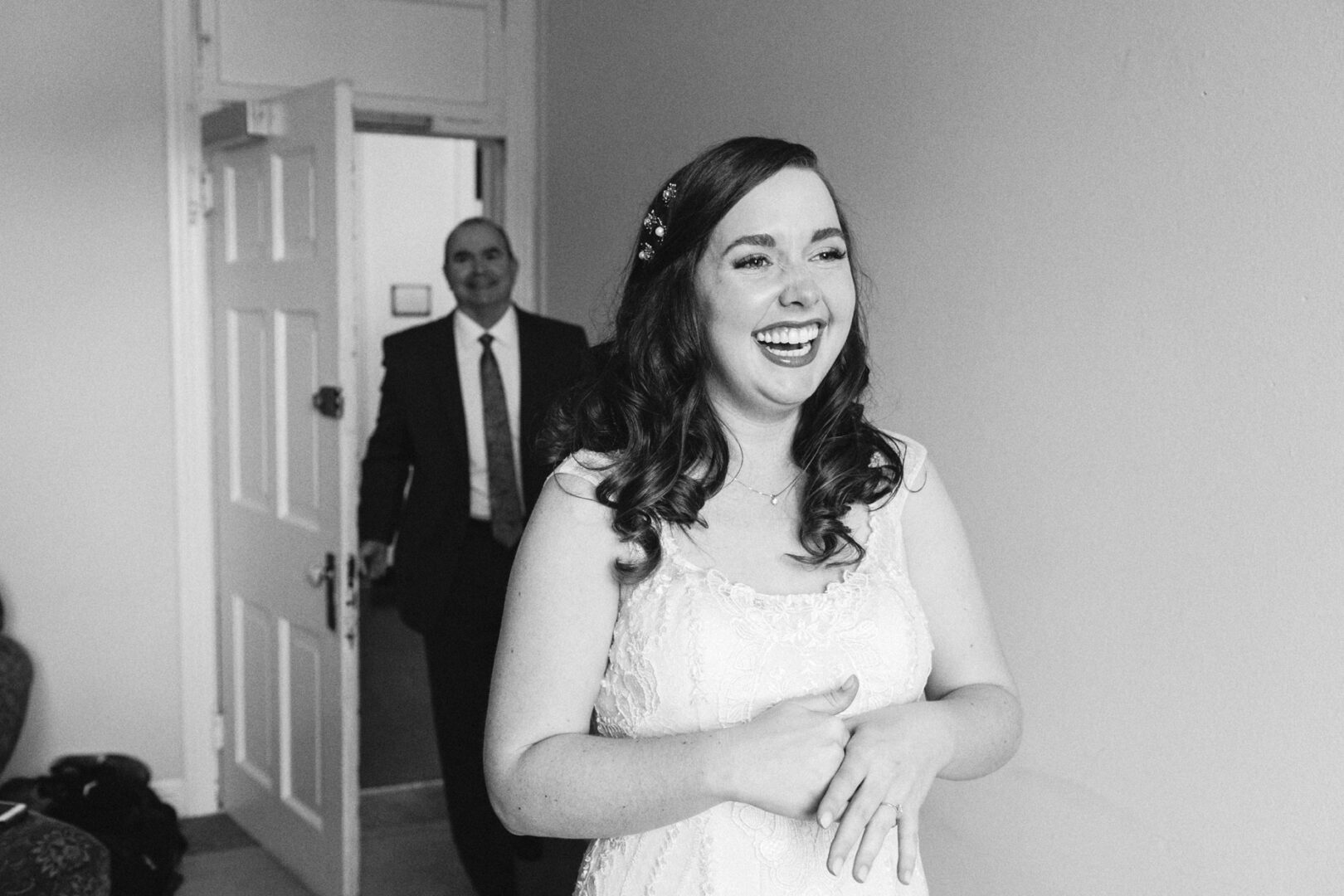 A bride in a white dress smiles joyfully as a man in a suit enters the room through an open door, capturing the charm of a Baldwin School wedding.