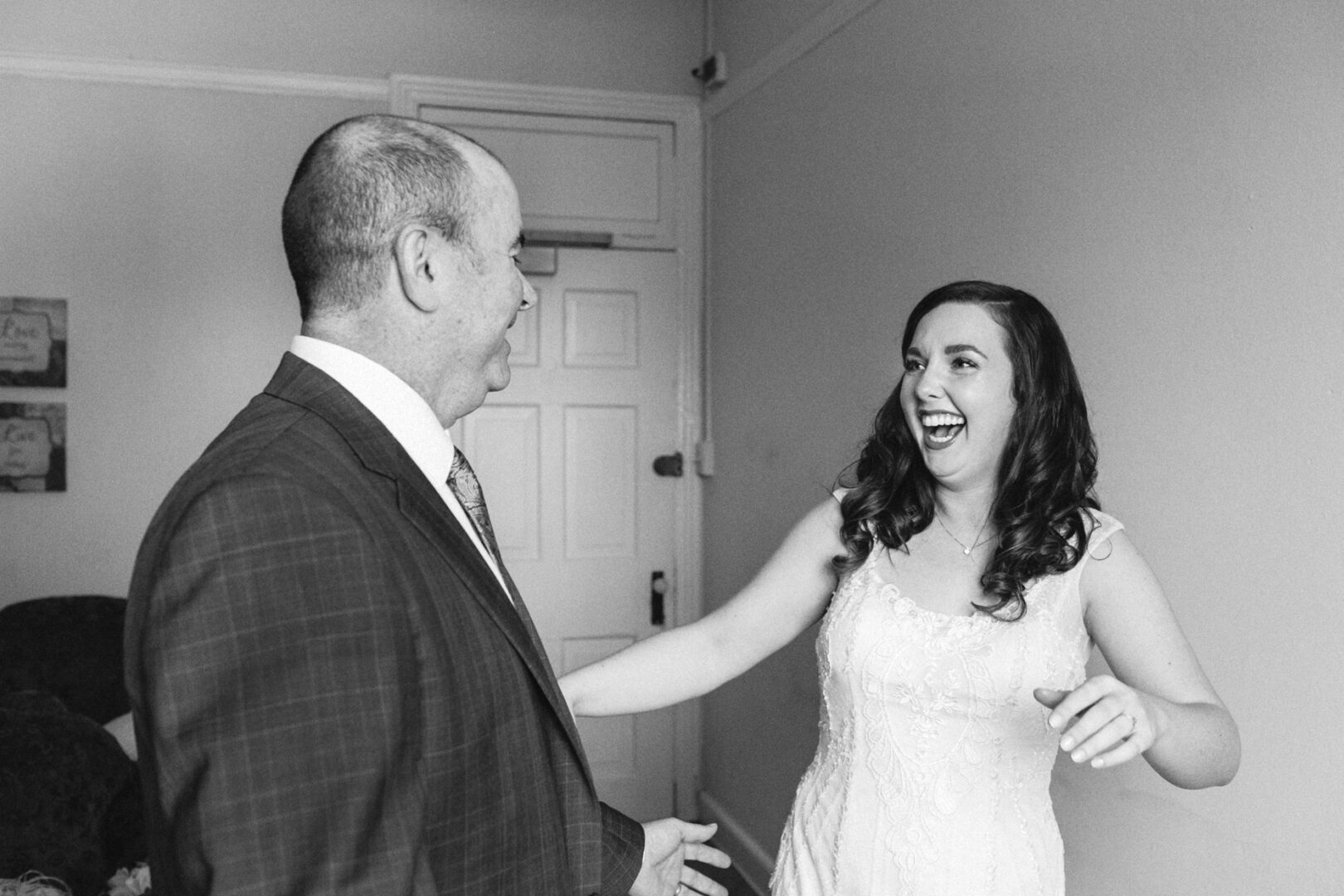 In a charming room at Baldwin School, a bride in a wedding dress joyfully greets a man in a suit, their smiles reflecting the joy of the wedding day.