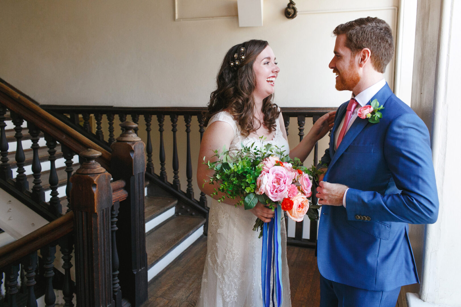 The bride, radiant in her white dress, and the groom, dapper in his blue suit with a pink tie, stand smiling near a wooden staircase at the picturesque Baldwin School wedding.