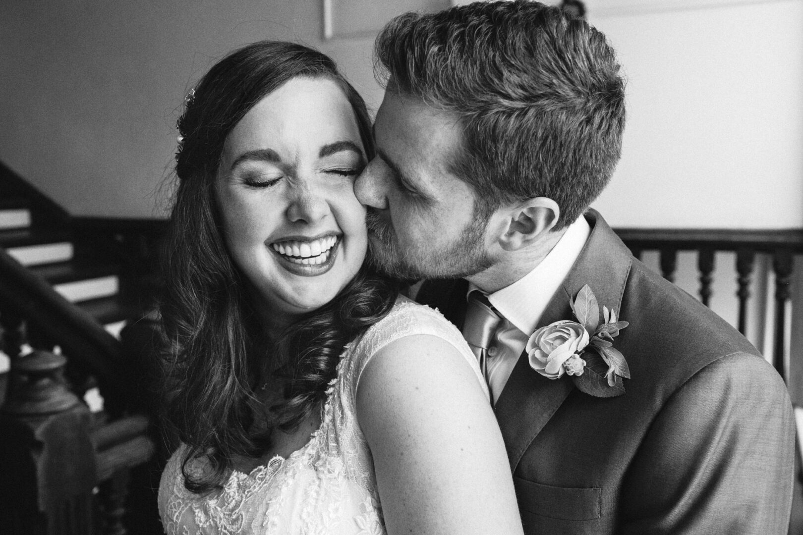 A couple dressed in wedding attire share a joyful moment on the staircase of Baldwin School. The man kisses the woman on the cheek, and they both smile. Black and white photo.