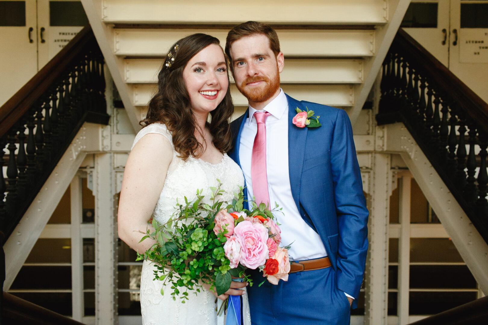 A bride and groom stand together smiling on the elegant staircase of Baldwin School. The bride holds a bouquet, and the groom wears a blue suit with a pink tie, both radiating joy on their wedding day.