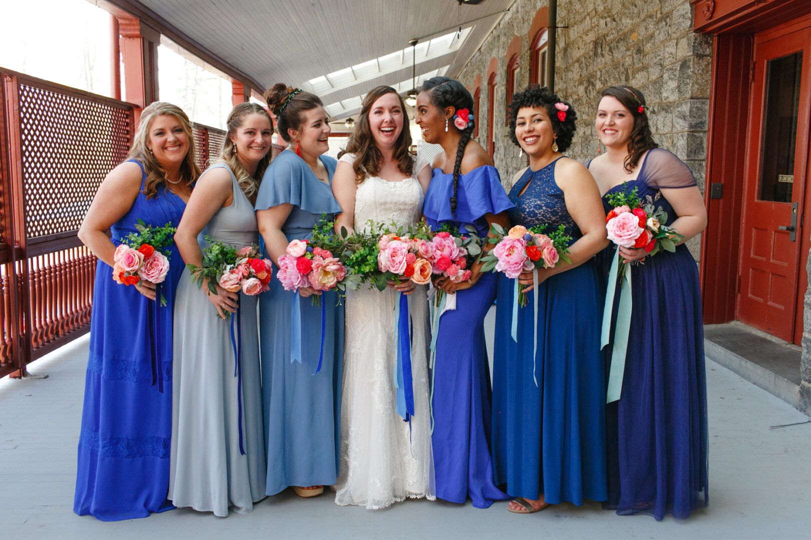 At a charming Baldwin School wedding, the bride in her white dress stands with her bridesmaids in elegant blue, each holding bouquets of pink and white flowers as they line up under the covered walkway.