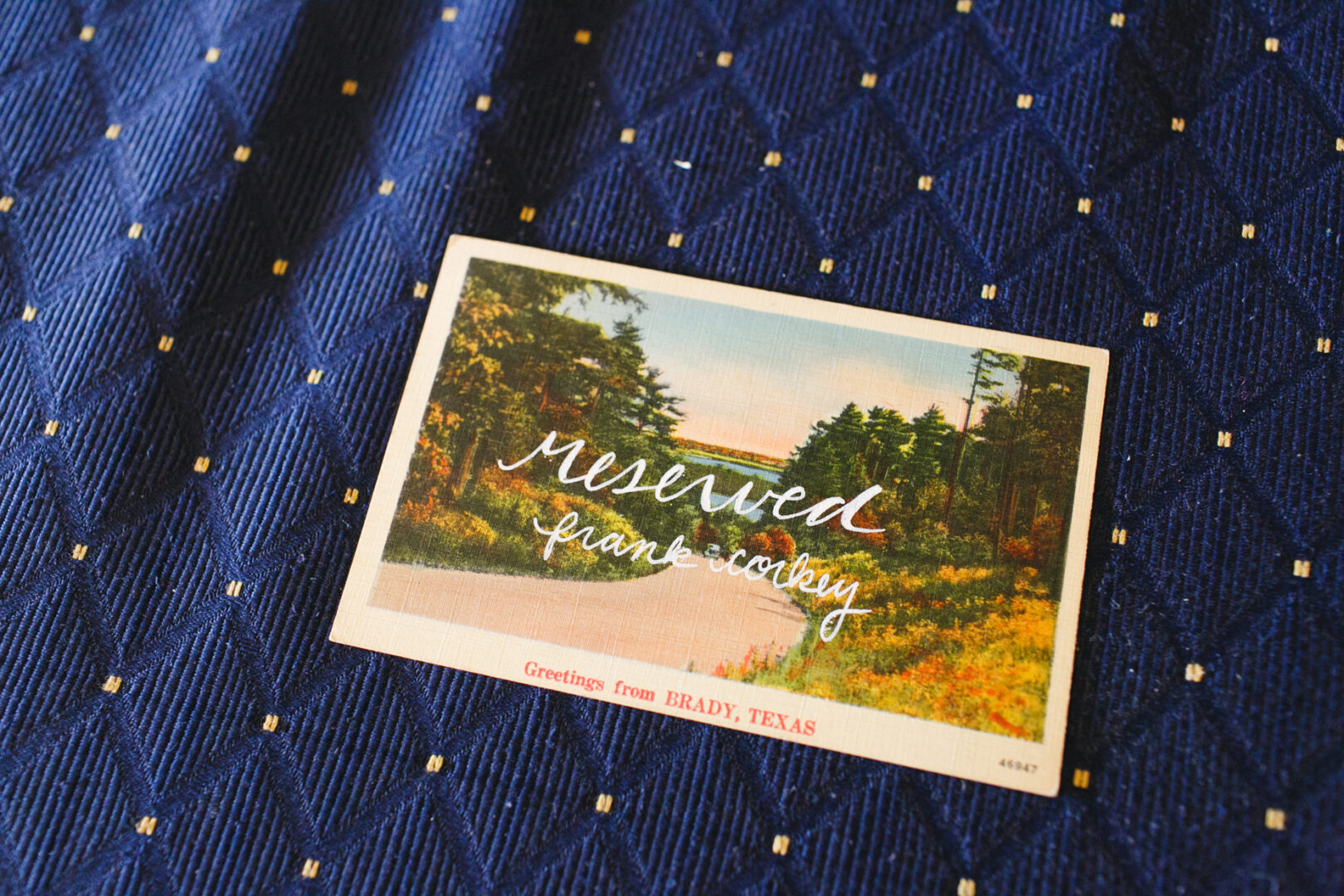 On a postcard with a scenic view, the words "reserved" and "Frank Conkey" elegantly sit atop a patterned blue fabric. Below, in charming script, it reads "Greetings from Brady, Texas," reminiscent of an invite to a Baldwin School Wedding.