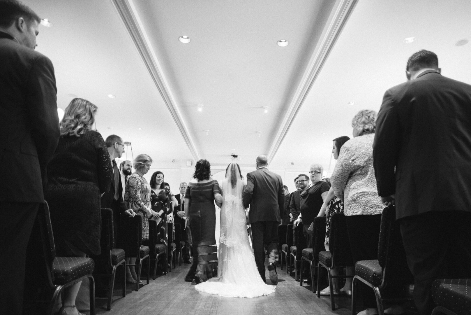 At the Baldwin School Wedding, the bride and groom gracefully walk down the aisle, surrounded by standing guests in a beautifully decorated hall.