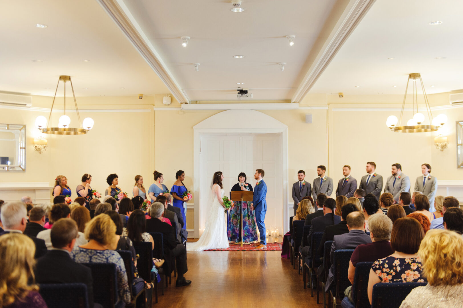 A Baldwin School Wedding unfolds as the couple stands at the altar with the officiant between them. Guests are seated, and the wedding party lines up on both sides in a room elegantly decorated.