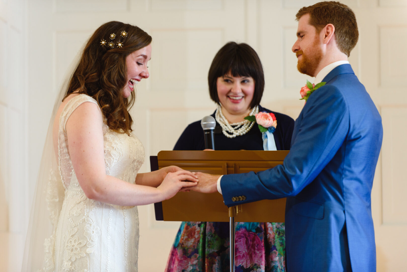 At a Baldwin School wedding, the bride and groom exchange rings, while a woman stands between them with a microphone to officiate the heartfelt ceremony.