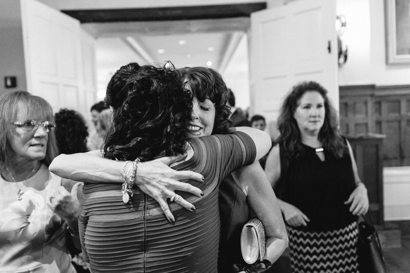 Two women warmly embrace in the foreground of a room at what appears to be a Baldwin School event. Others stand nearby, observing or engaged in conversation, reminiscent of a joyous wedding celebration.