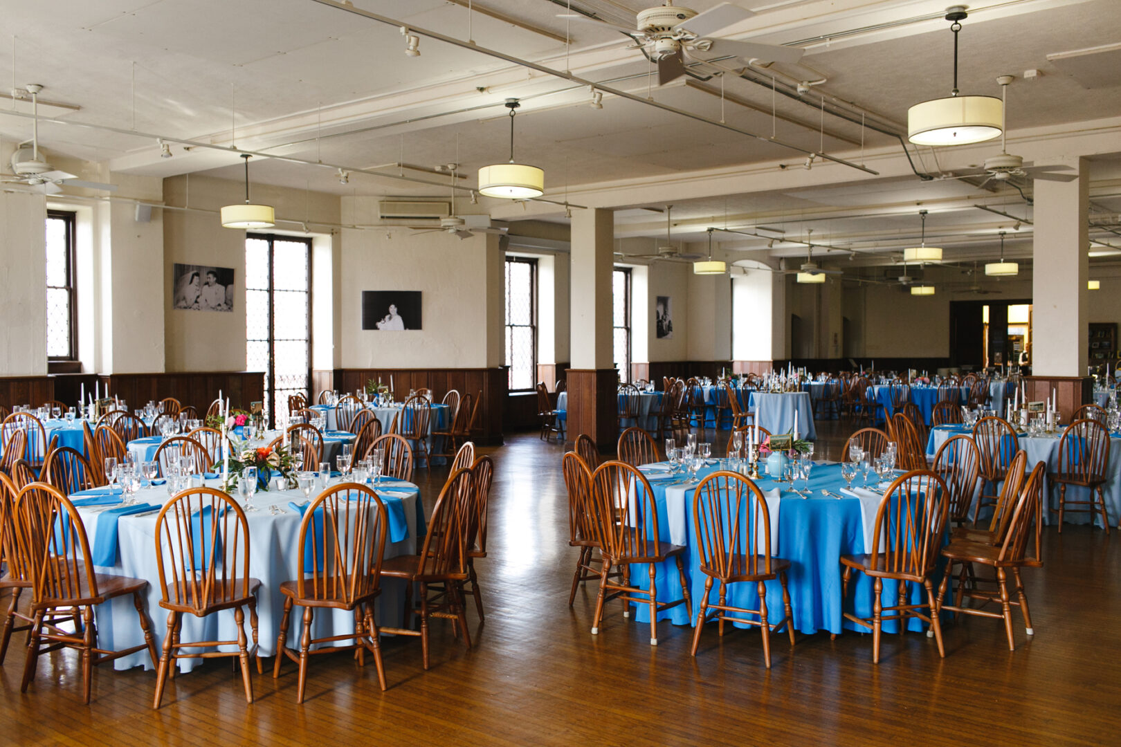 A banquet hall with round tables draped in blue tablecloths hosts a Baldwin School wedding. Each table is elegantly set with dinnerware, glassware, and floral centerpieces, as wooden chairs encircle them.