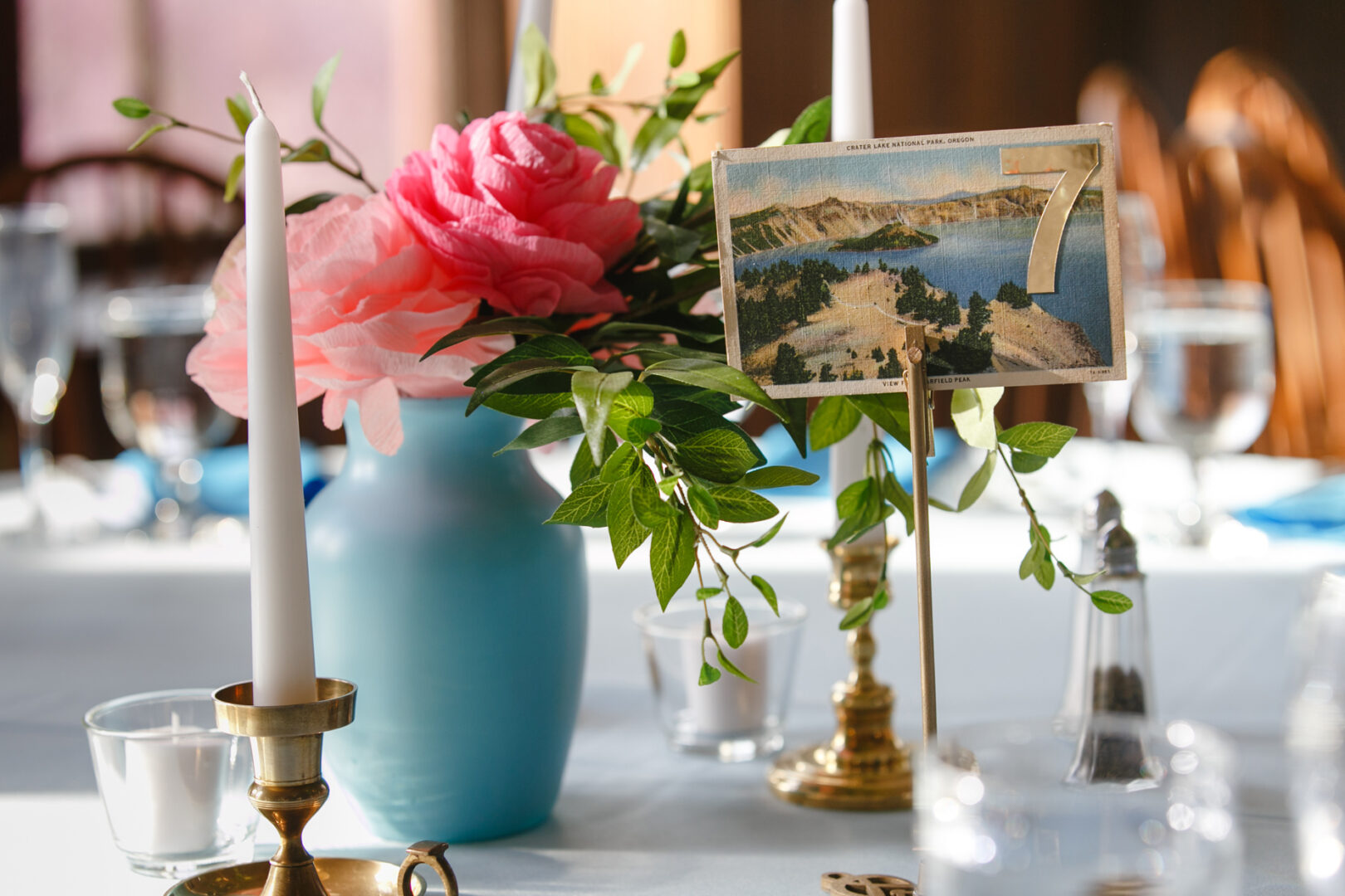 A beautiful table setting at the Baldwin School wedding features a blue vase filled with pink flowers and lush greenery. A card displaying the number "7" with a landscape image sits elegantly in a gold holder, surrounded by flickering candles and sparkling glassware on a pristine white tablecloth.