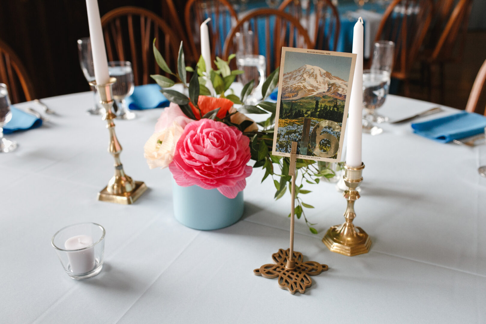 A Baldwin School Wedding-themed table setting features a floral centerpiece, elegant candlesticks, and a photo holder showcasing a mountain postcard.