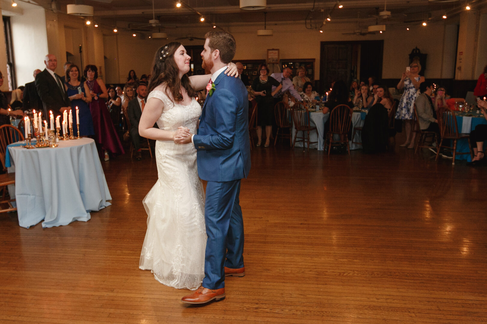 At the Baldwin School Wedding, a bride in a white dress and a groom in a blue suit gracefully dance together, their movements enchanting the guests seated around them in the spacious room.