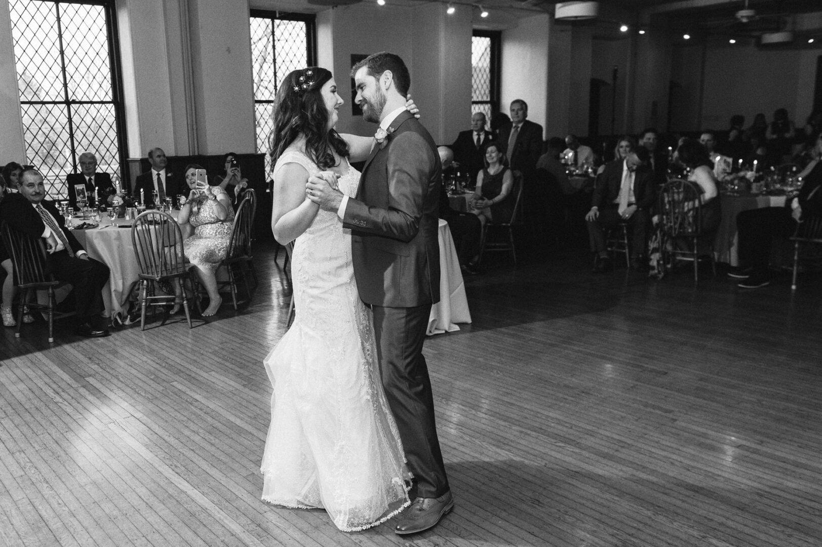 At a Baldwin School wedding, a bride and groom share their first dance in the elegant reception hall. Guests sit at tables around the room, watching in admiration as the classic atmosphere adds a timeless touch to their special moment.
