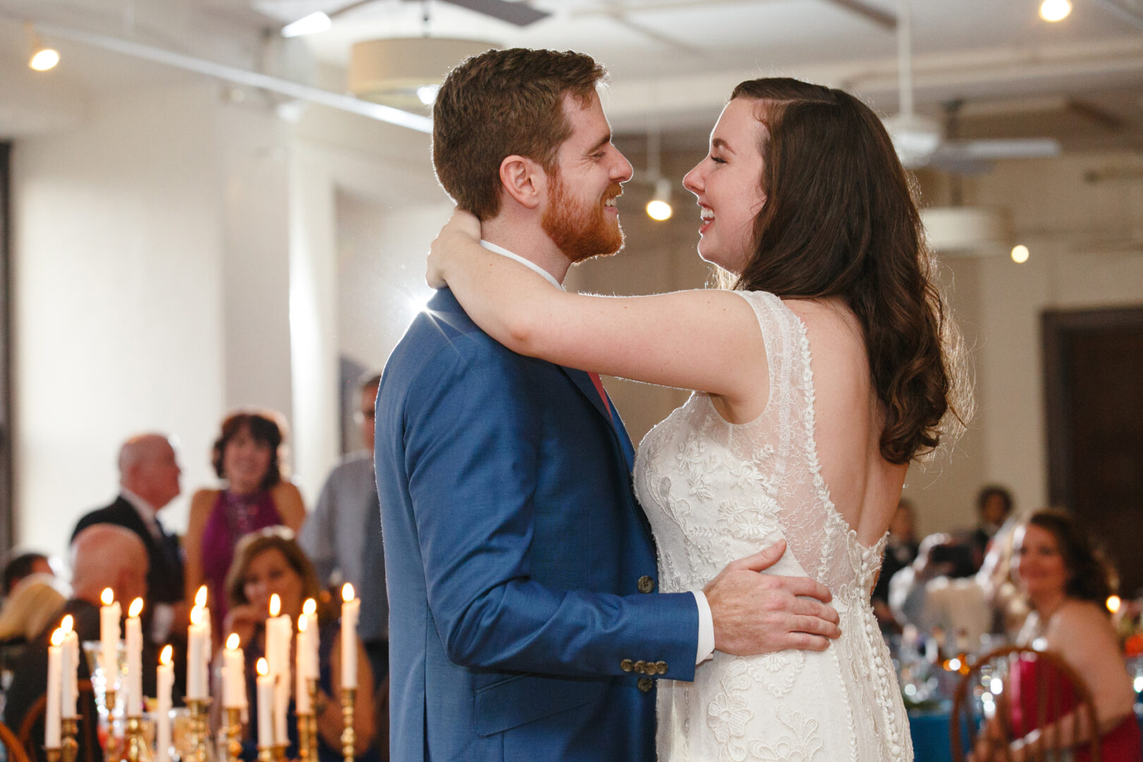A couple, dressed in a blue suit and white lace gown, dance together in the warmly lit Baldwin School Wedding venue. Guests are seated in the background.