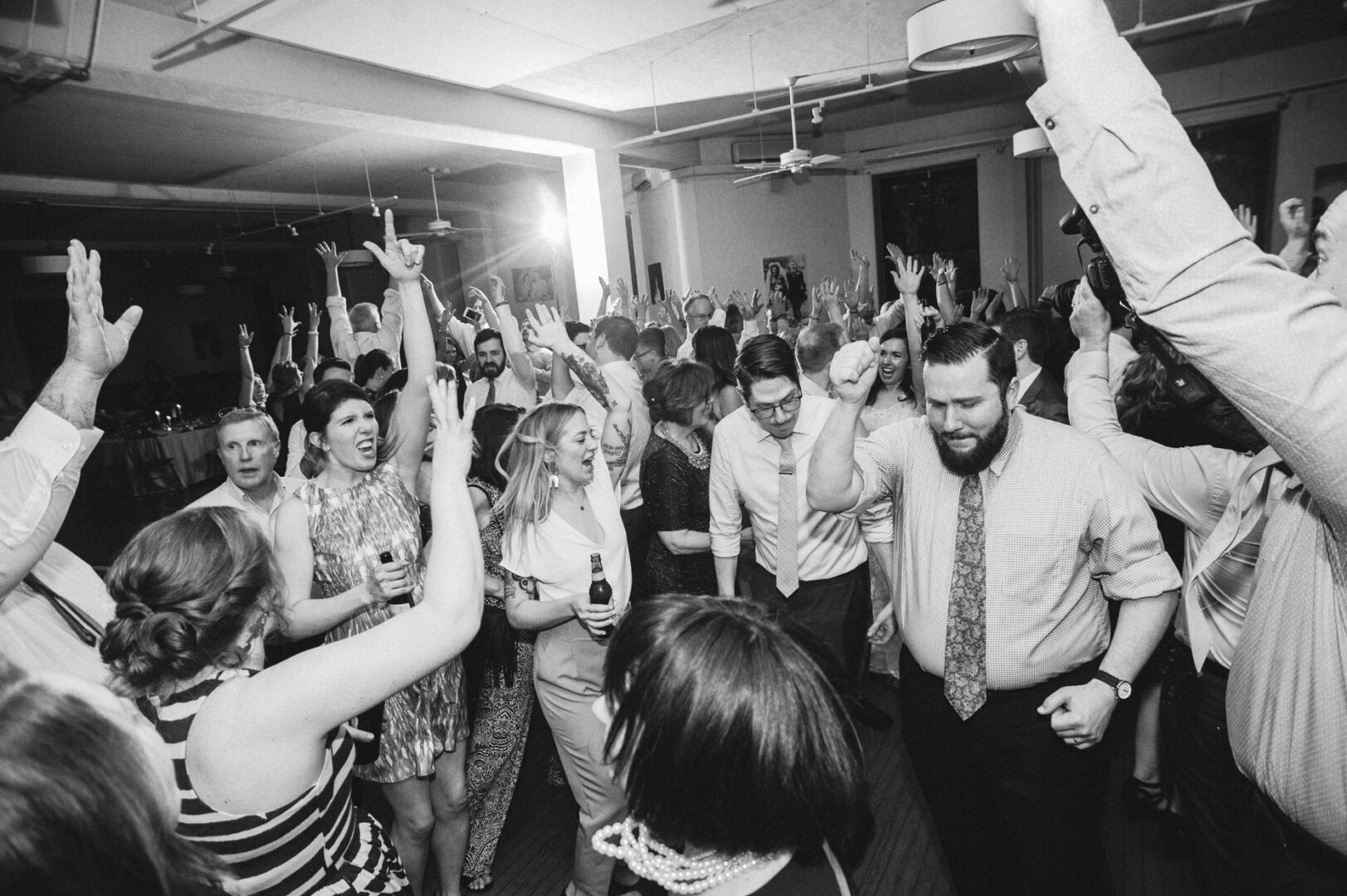 At the Baldwin School wedding, a lively crowd dressed in formal attire fills the indoor venue. With drinks in hand, they dance exuberantly with raised arms, celebrating the joyous occasion.