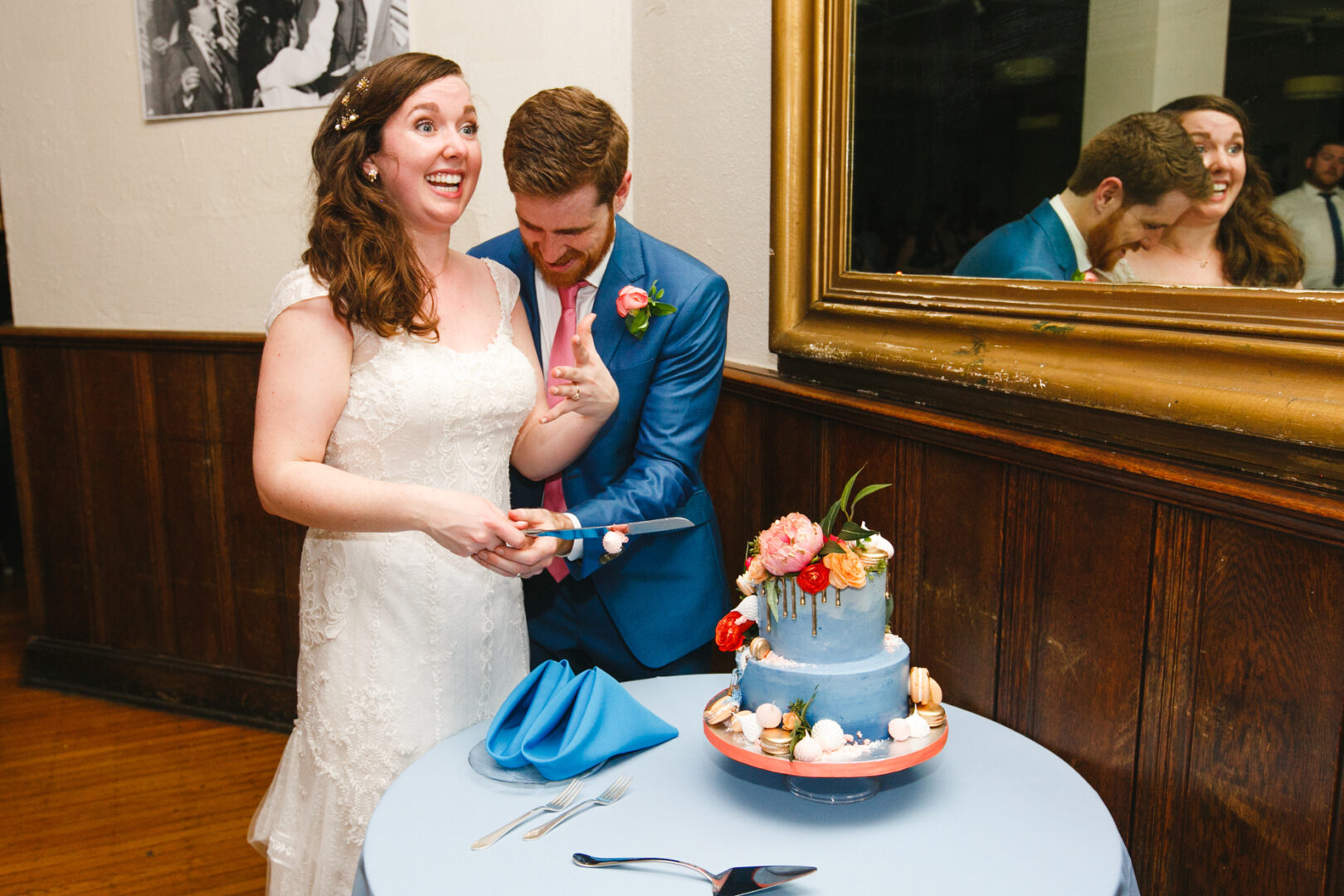 A bride and groom in wedding attire prepare to cut a blue two-tiered cake adorned with flowers, standing beside a large mirror in the wood-paneled room of the Baldwin School. A photo hangs on the wall behind them, capturing the essence of this special Baldwin School wedding.