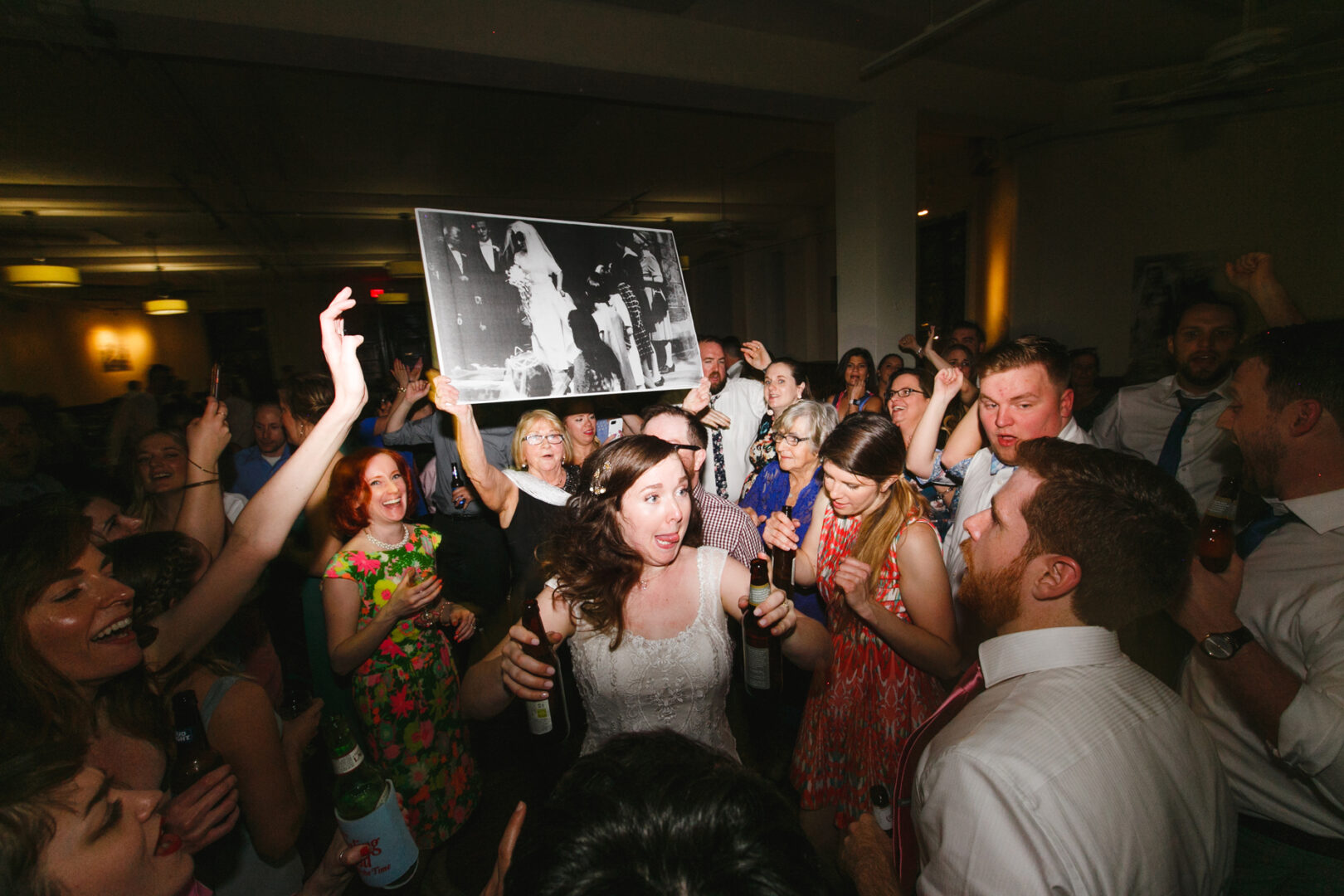 A crowd of people cheer and celebrate at a Baldwin School wedding. A woman in focus holds a black and white photo, capturing a cherished memory. Many hold drinks, and the lighting suggests an indoor setting filled with joy and laughter.