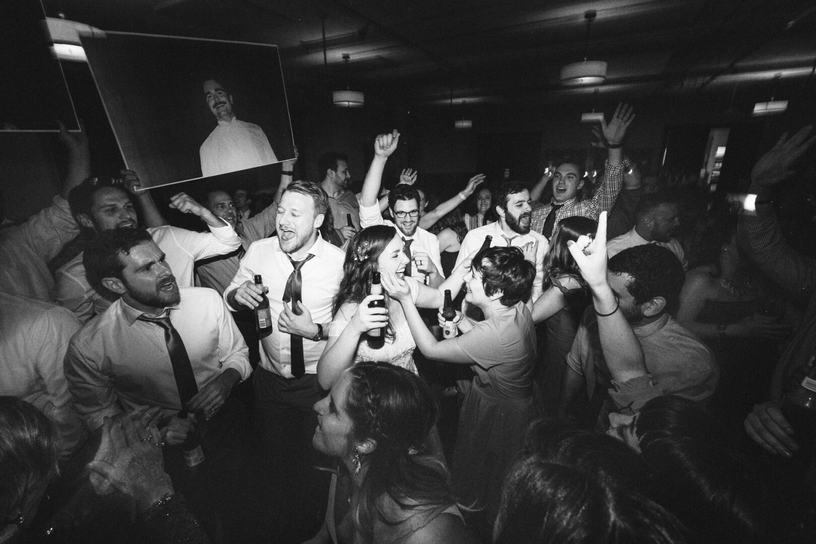 A group of people at a lively Baldwin School wedding, many holding drinks and cheering, with black-and-white decor and a cutout photo of a smiling man visible in the background.