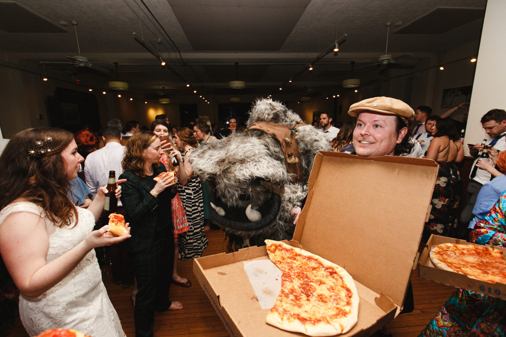 At a Baldwin School wedding, a man in a cap offers pizza from a box at the lively celebration, while another person in a furry costume entertains guests. Laughter fills the air as everyone savors their pizza slices, creating an unforgettable atmosphere.