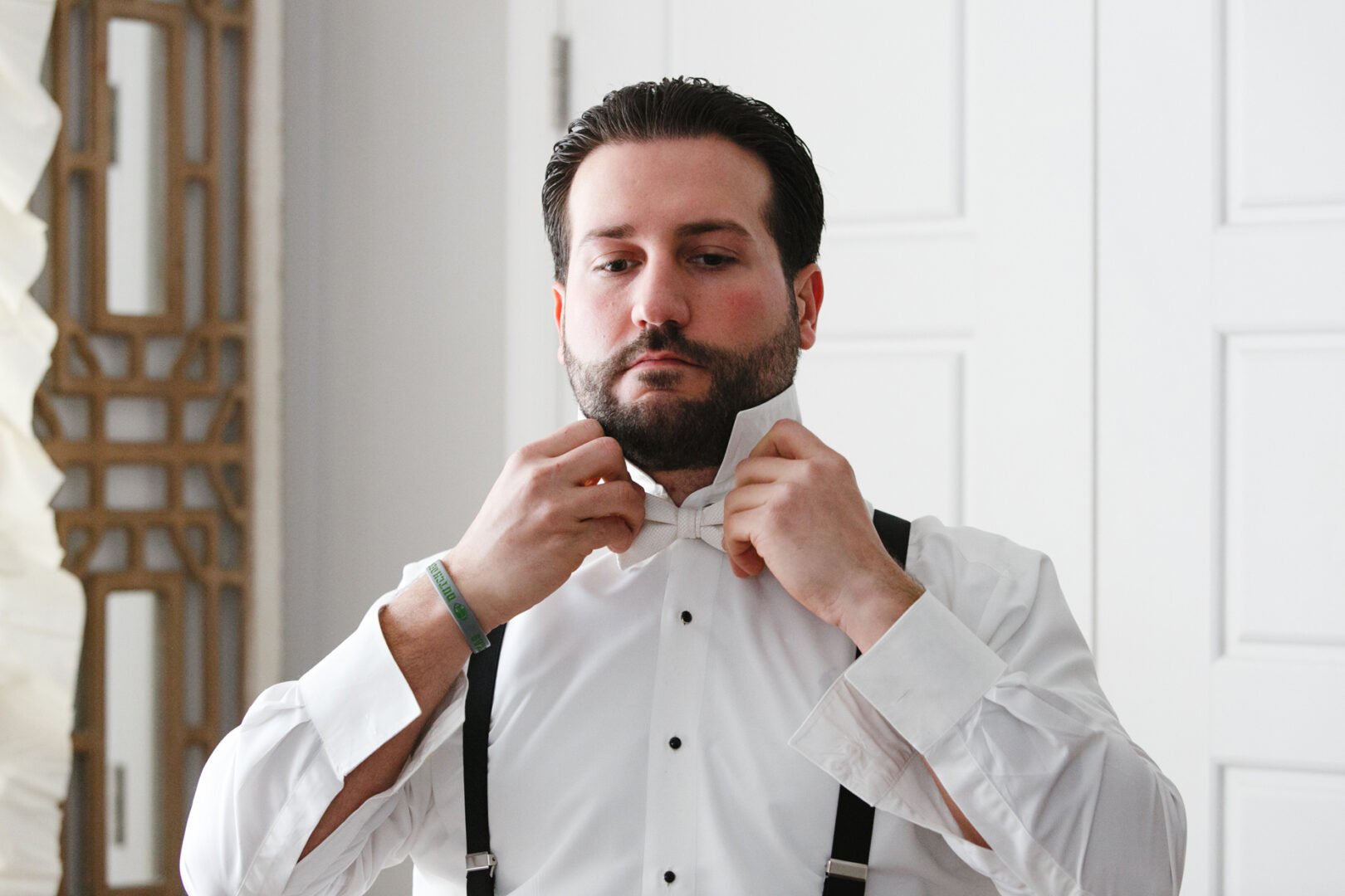 Man in a white dress shirt adjusts his bow tie in front of a white door, the room echoing with memories of a Crystal Tea Room wedding, his suspenders elegantly hanging by his side.