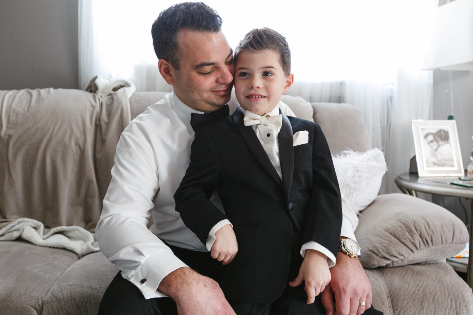 Man and young boy in formal suits sit on a sofa, beaming amid the elegant ambiance of a Crystal Tea Room wedding. A framed photo graces the side table, capturing the essence of their joyous day.
