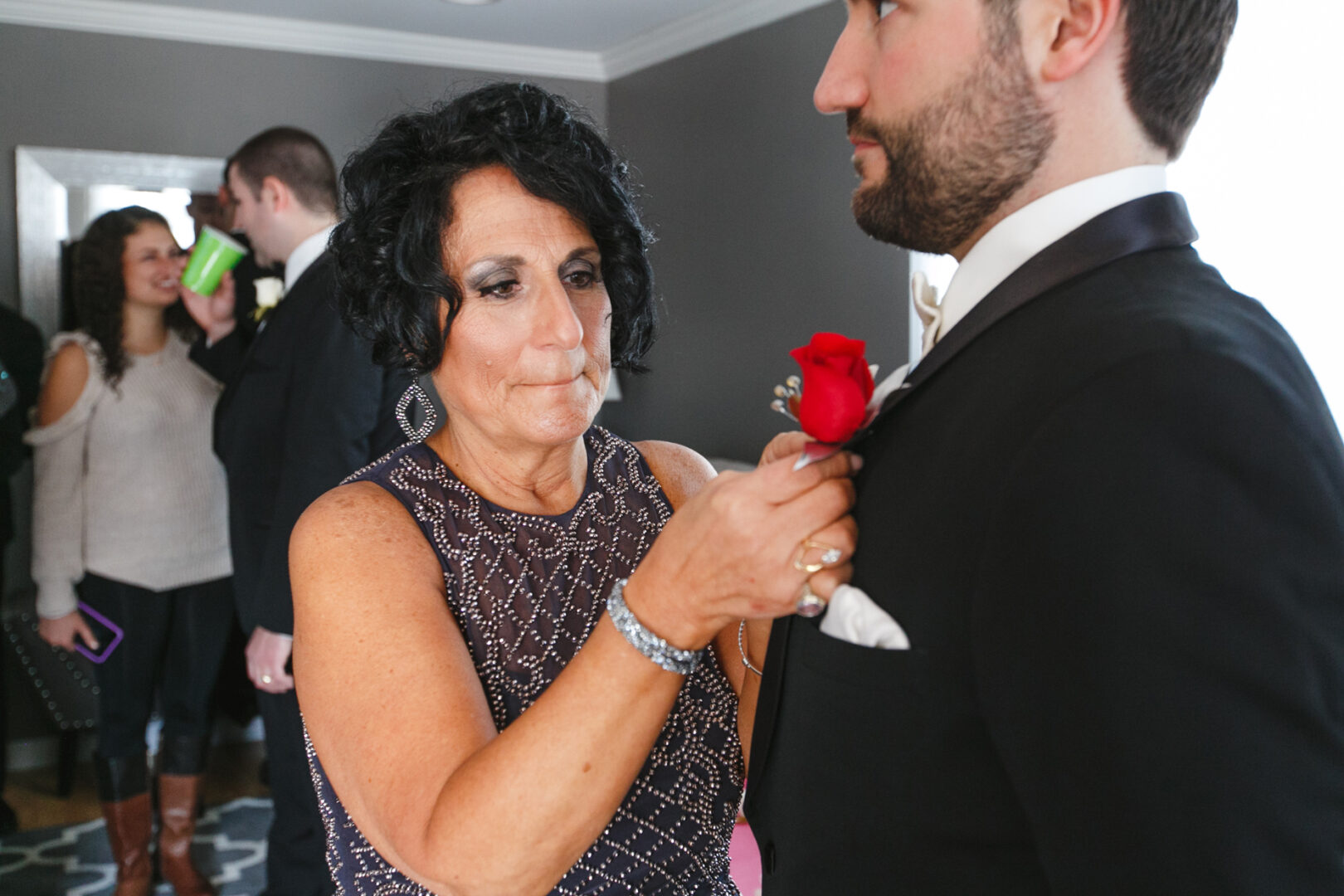 At a Crystal Tea Room wedding, a woman adjusts a boutonniere on a man's tuxedo. In the background, guests mingle, with one holding a green cup.