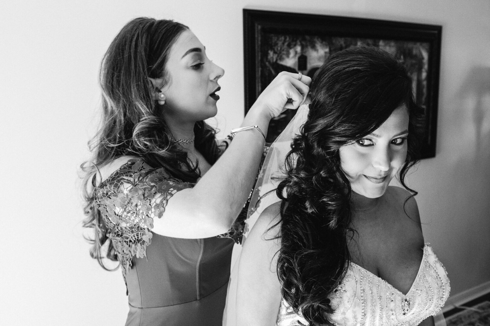 In the elegant setting reminiscent of a Crystal Tea Room wedding, a woman in a dress expertly styles another woman's hair as she dons a graceful gown. They are in a room adorned with a framed picture on the wall.