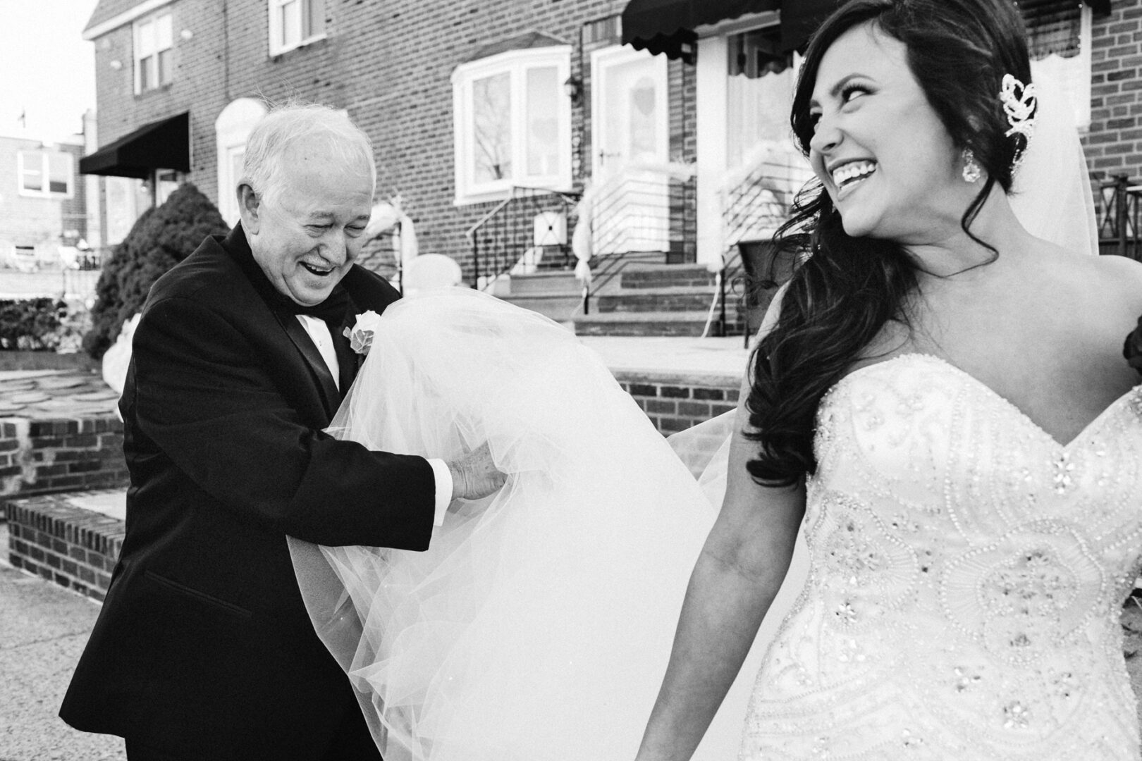 A bride smiles broadly as an older man in a suit holds up her dress, their laughter echoing against the brick buildings. It's a charming moment from their Crystal Tea Room wedding.