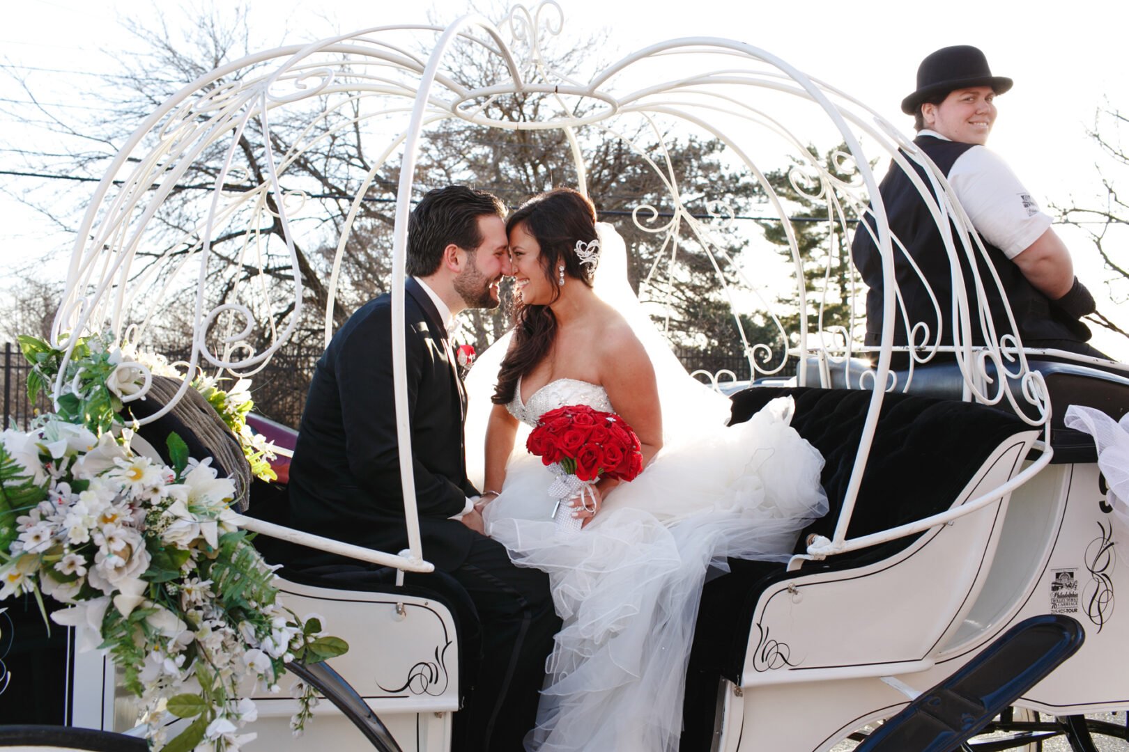 A couple in wedding attire sits inside a white carriage, the bride clutching red roses, as if they are on their way to an enchanting Crystal Tea Room wedding. A driver in a vest and bowler hat is seated at the front.
