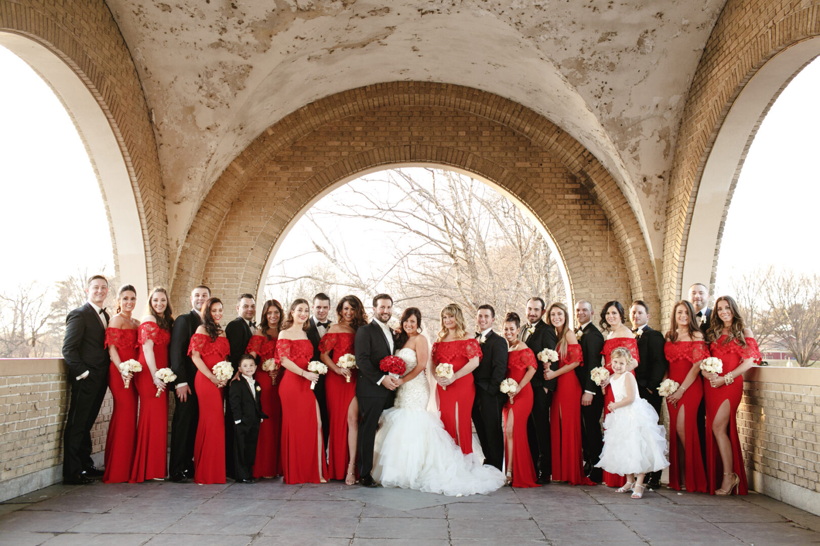 A large wedding party in elegant attire stands under an arched brick structure at the Crystal Tea Room. Bridesmaids in red dresses, groomsmen in black suits, and two brides hold bouquets.
