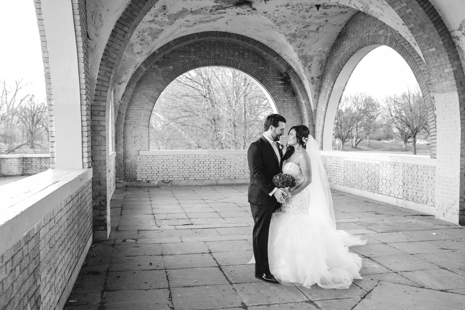 Bride and groom stand under an arched structure at the Crystal Tea Room, facing each other. The bride holds a bouquet, and both wear elegant wedding attire. Black and white image.