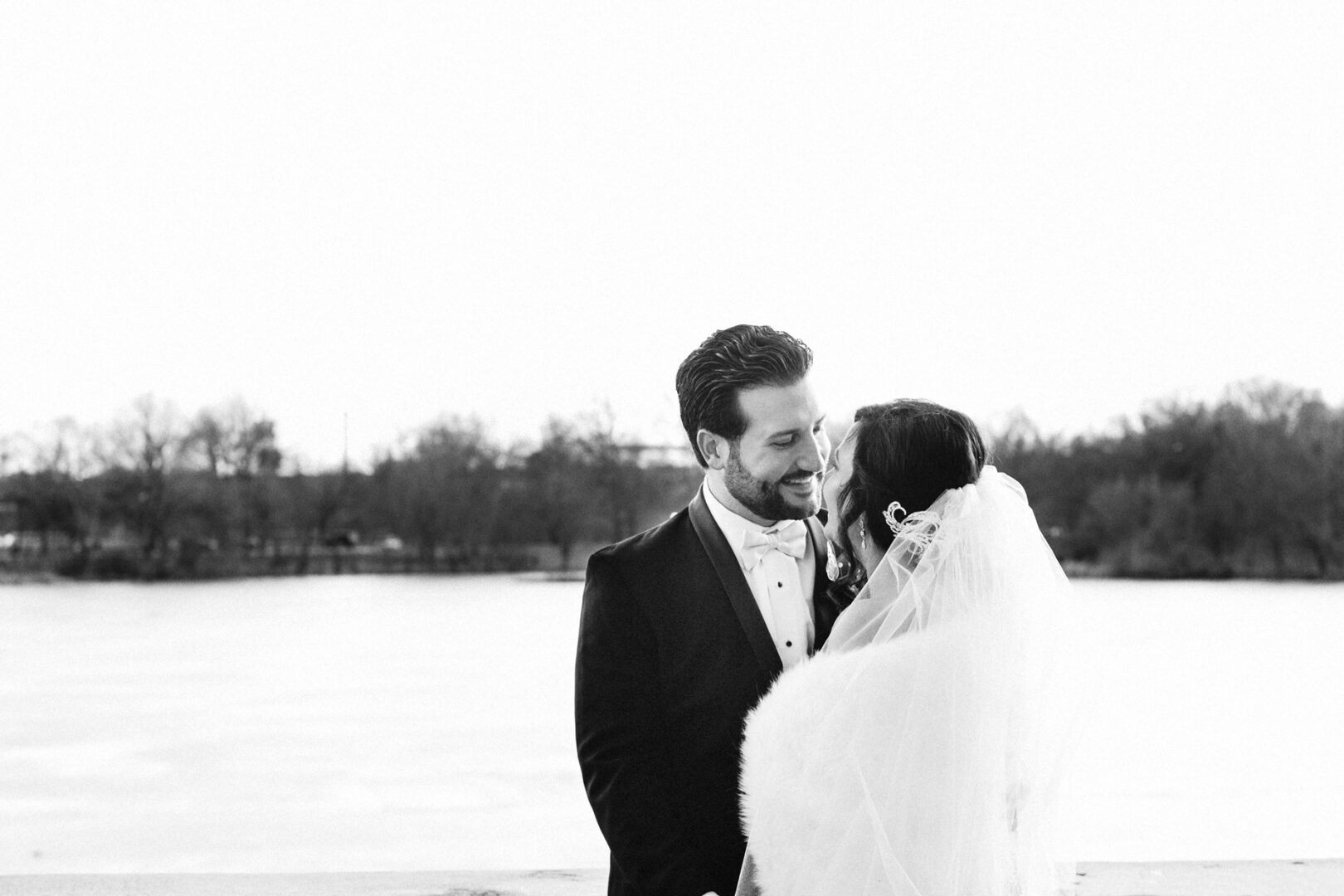 A couple in wedding attire, facing each other and smiling, stands outdoors near a body of water. The scene is monochrome, with trees visible in the background, evoking the timeless elegance of a Crystal Tea Room wedding.