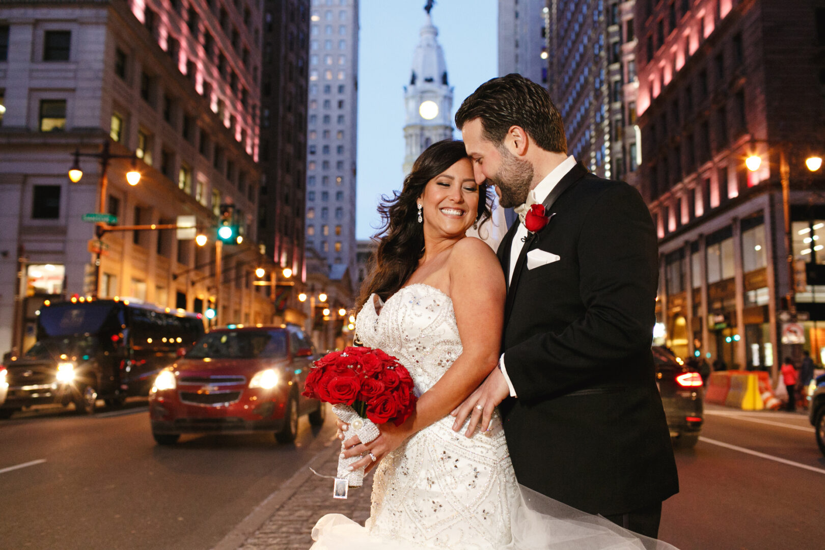 A bride and groom embrace on a city street at night, surrounded by the glow of lit buildings and vibrant traffic. The couple, fresh from their Crystal Tea Room wedding, share a tender moment as the bride holds a bouquet of red flowers.