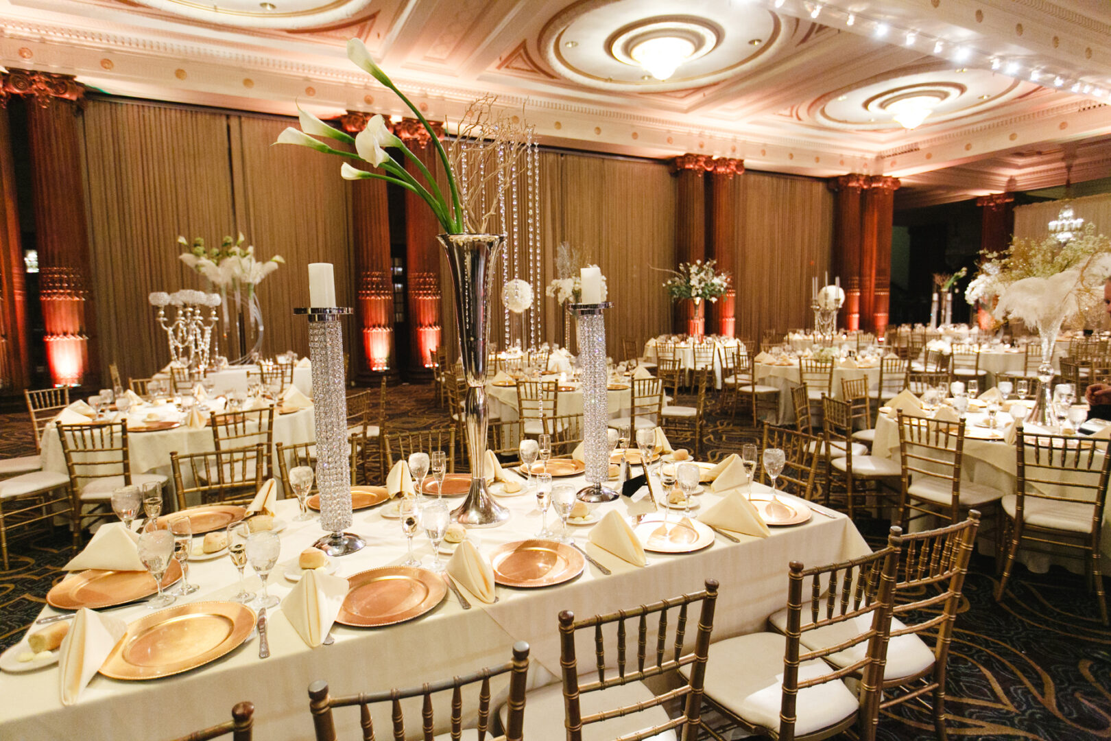 The elegant ballroom, perfect for a Crystal Tea Room wedding, features round tables adorned with white tablecloths and gold chairs. Tall floral centerpieces enhance the ambiance, while soft lighting creates a sophisticated atmosphere for your special day.
