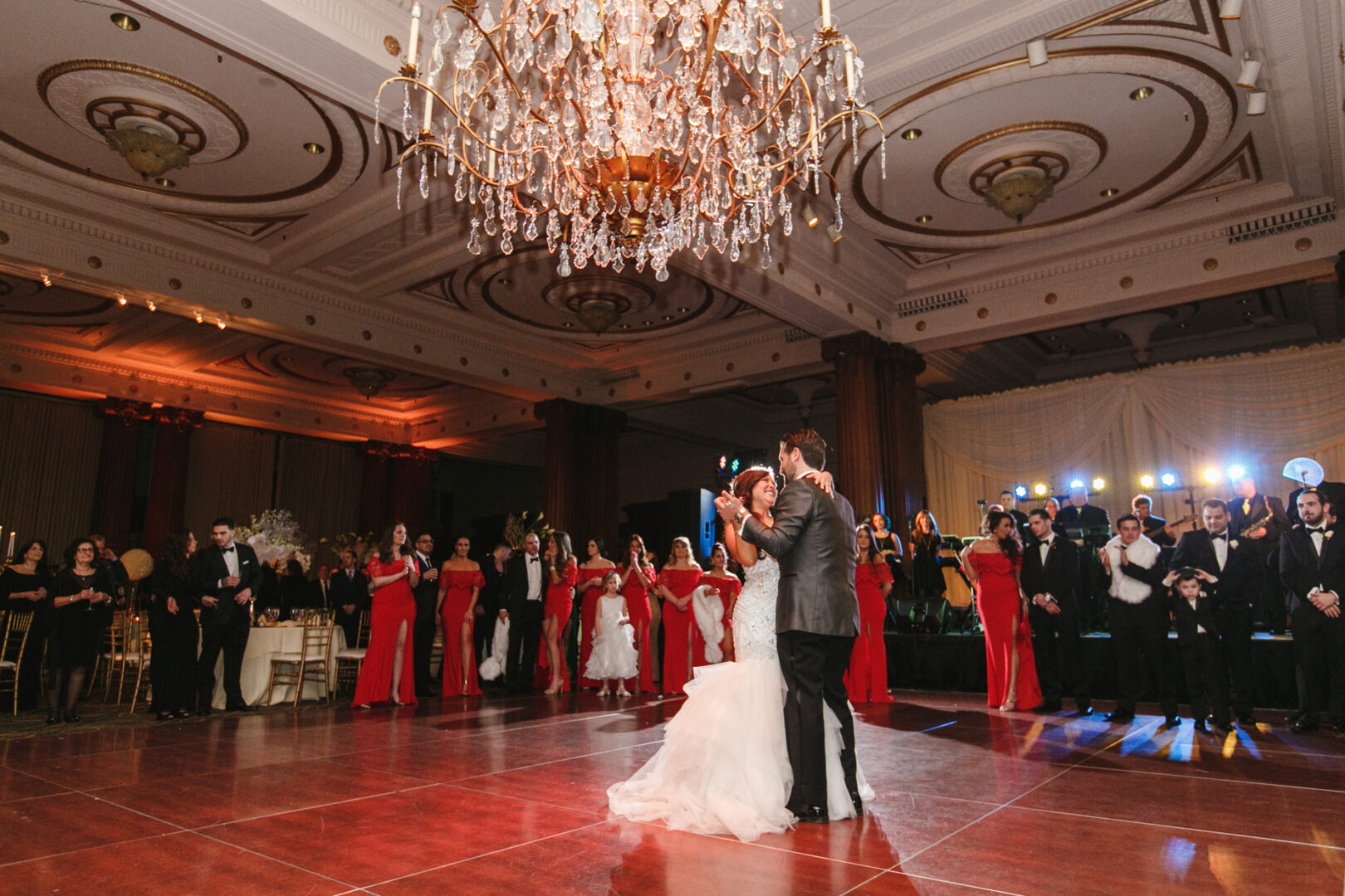 A couple dances under a grand chandelier in an elegant ballroom, reminiscent of a Crystal Tea Room wedding. Surrounded by guests, women in red dresses and men in formal attire watch the pair.