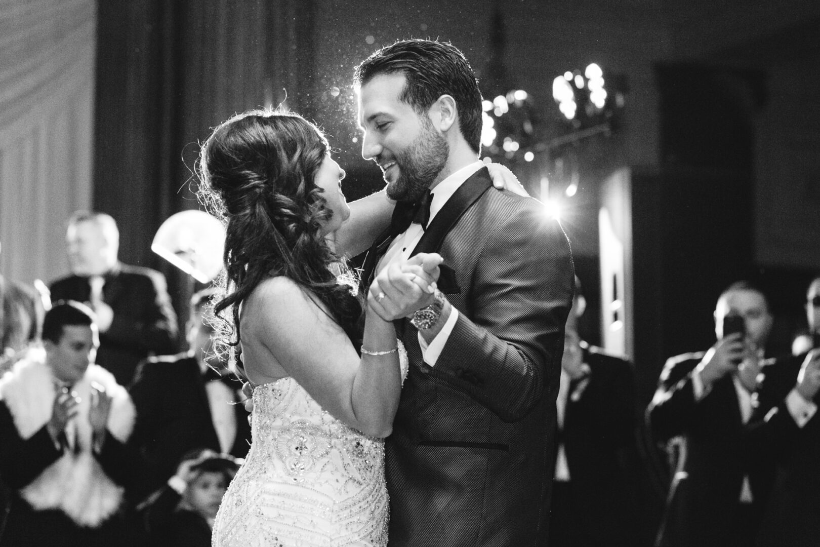 At a Crystal Tea Room wedding, a couple dances closely in formal attire, surrounded by applauding guests. The scene is black and white, with the focus gracefully capturing their shared smiles and joyous moment.