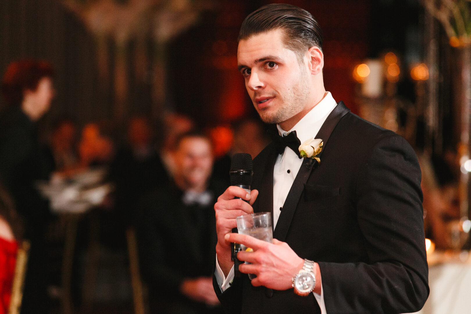 At a Crystal Tea Room wedding, a man in a tuxedo holds a microphone and a drink, addressing the attendees while the lively atmosphere buzzes in the background.