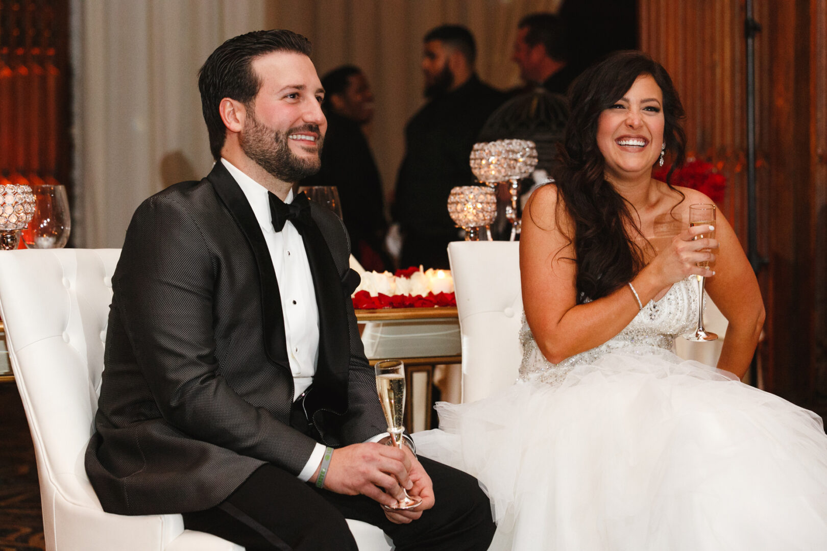 At the Crystal Tea Room wedding, the bride and groom, elegantly dressed, are seated together with joyful smiles as they raise their glasses in celebration at the reception.