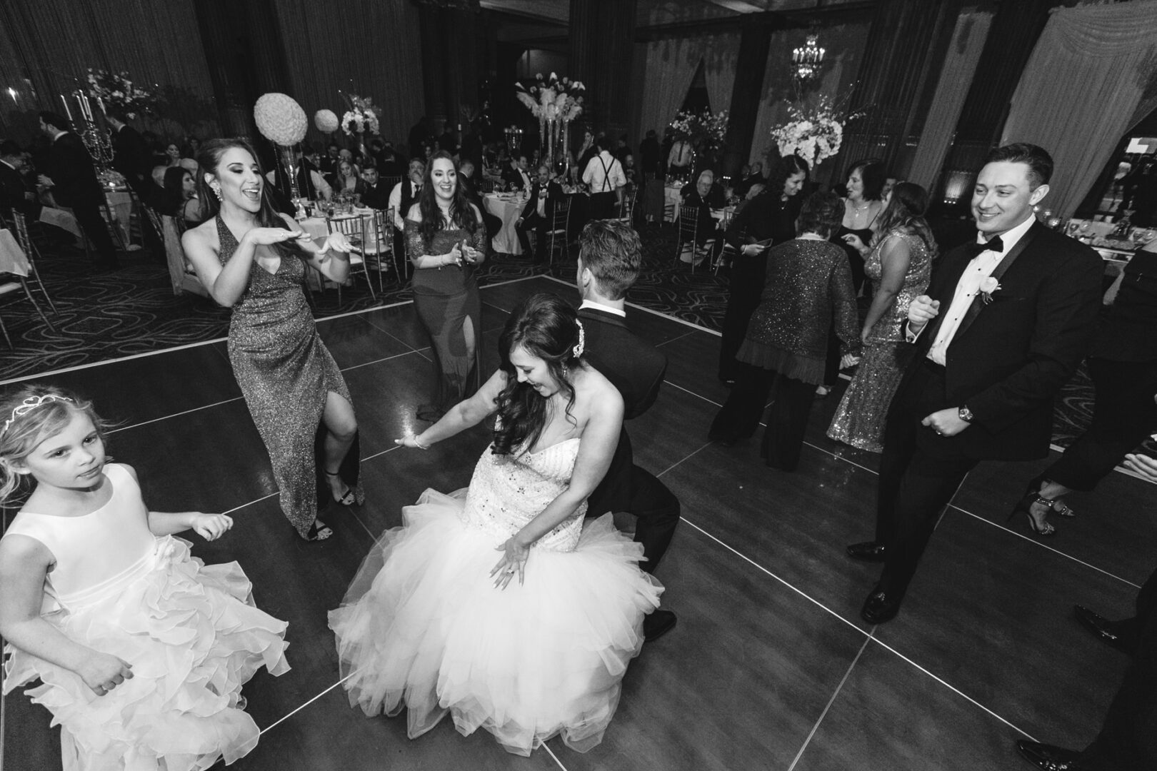 At the Crystal Tea Room wedding, a bride and groom dance energetically on the ballroom floor. Guests, including a young girl, watch and join in. The scene is lively with formal attire and elegant decorations.