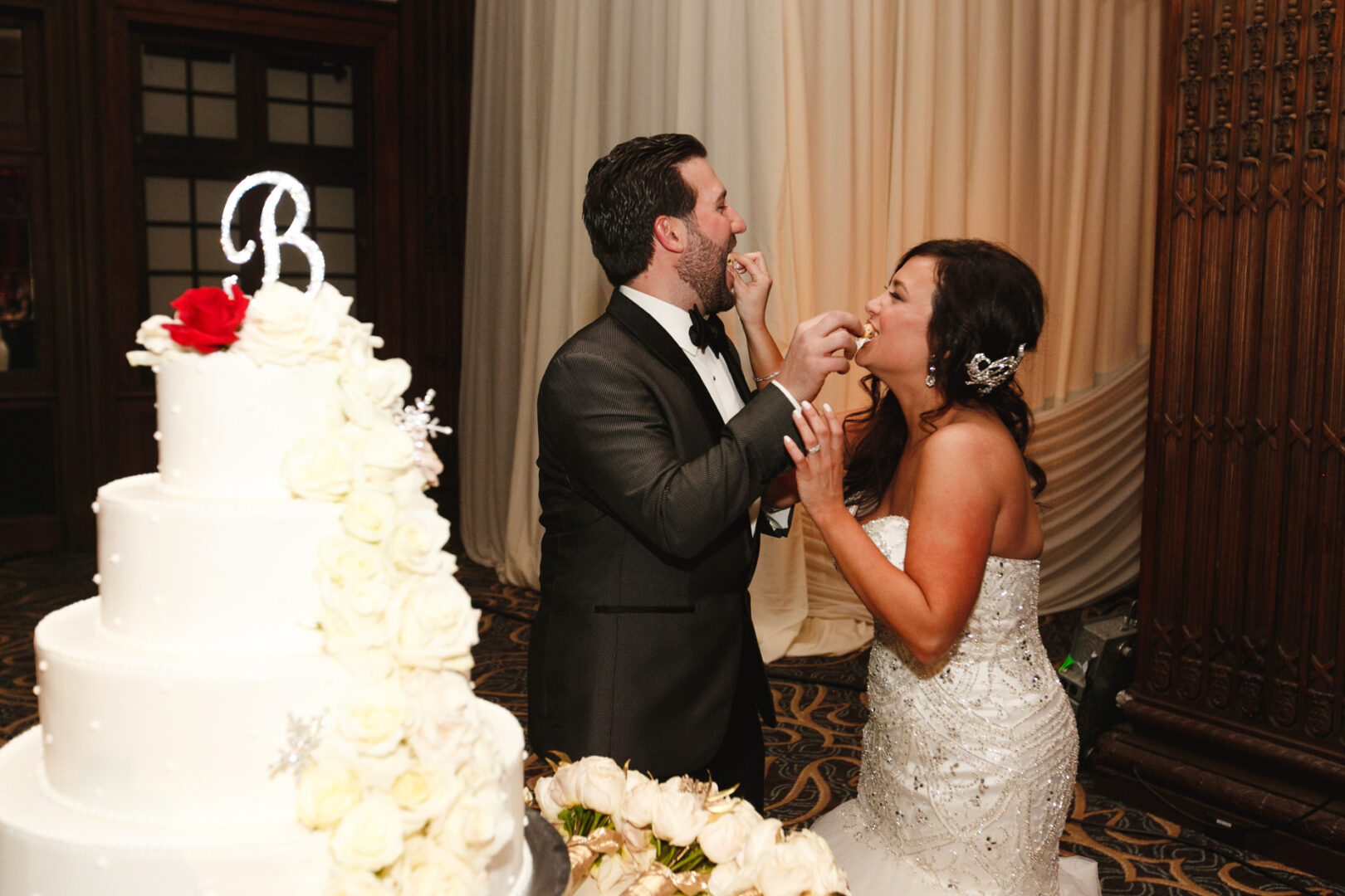 At their Crystal Tea Room wedding, the couple lovingly fed each other cake beside a tiered masterpiece adorned with white roses and a "B" topper.