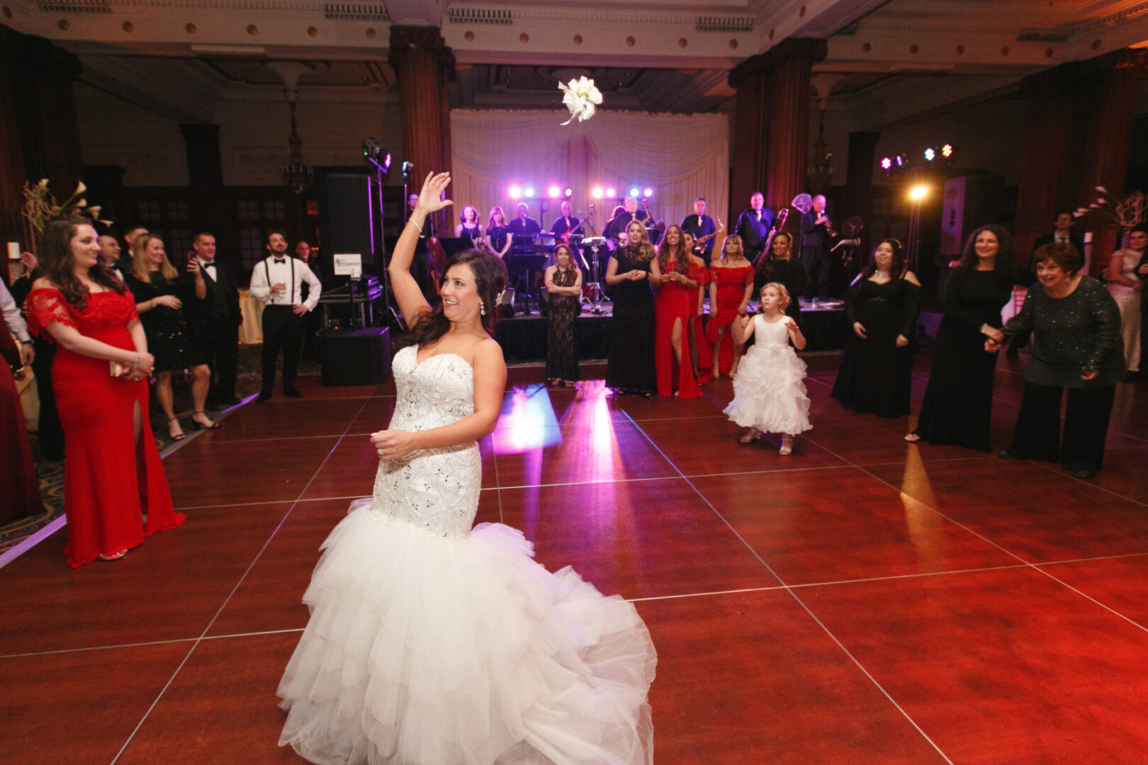 The bride, in a stunning white gown, elegantly tosses her bouquet in the grand Crystal Tea Room ballroom, with guests in formal attire watching eagerly.