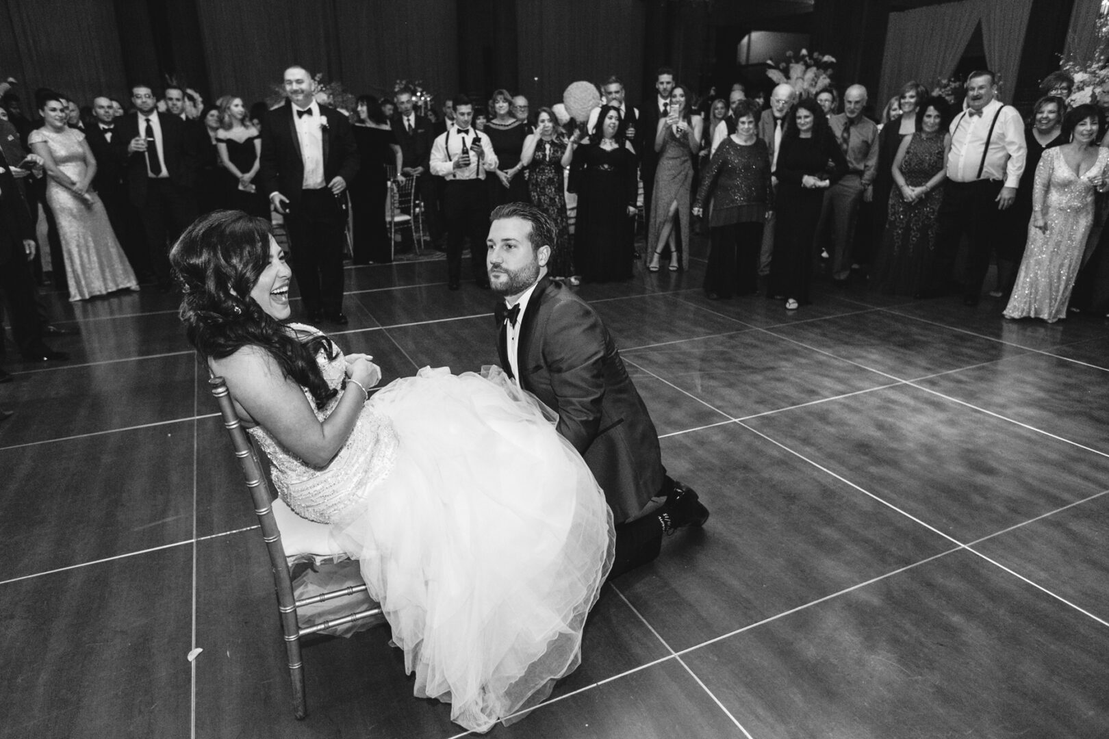 At a stunning Crystal Tea Room wedding, the groom kneels in front of his seated bride on the dance floor, surrounded by elegantly dressed guests.